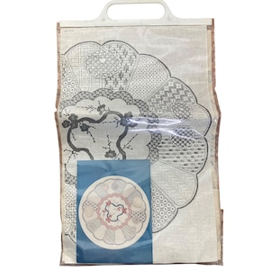 Puede incluir: Un artículo textil beige con un diseño floral, empaquetado en una bolsa de plástico transparente con un asa blanca. El diseño presenta un gran patrón semicircular con varios motivos geométricos y florales en gris y negro. Se ve un diseño circular más pequeño.