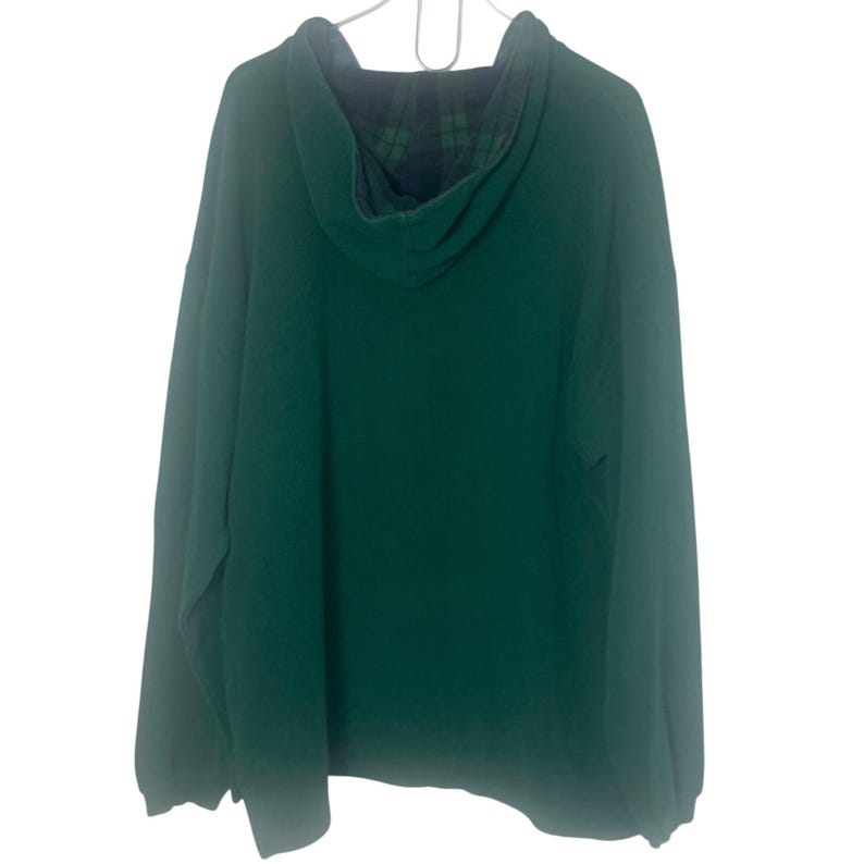 May include: A long-sleeve pullover hoodie in a solid dark green color. The garment features a hood lined with a dark blue and green plaid pattern. The hoodie is displayed on a white hanger against a plain white background, showing the back view.