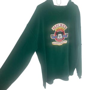 May include: A dark green long-sleeve hooded sweatshirt featuring an embroidered graphic on the chest. The design includes the text "MICKEY" and "EST. 1928" above a circular emblem with a Mickey Mouse face, the words "VERITAS" and "ARTIS", and additional text.