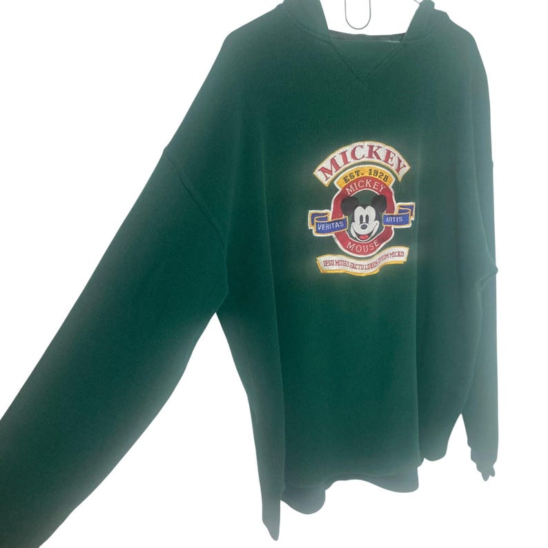 May include: A dark green long-sleeve hooded sweatshirt featuring an embroidered graphic on the chest. The design includes the text "MICKEY" and "EST. 1928" above a circular emblem with a Mickey Mouse face, the words "VERITAS" and "ARTIS", and additional text.