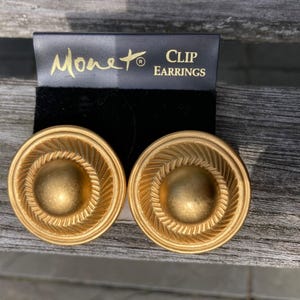 Könnte beinhalten: Ein Paar goldfarbene Clip-Ohrringe mit kreisförmigem Design. Die Ohrringe haben eine zentrale, erhabene Kuppel, die von strukturierten, konzentrischen Ringen umgeben ist. Die Ohrringe werden auf einer schwarzen Karte mit dem "Monet"-Logo und dem Text "CLIP EARRINGS" präsentiert.