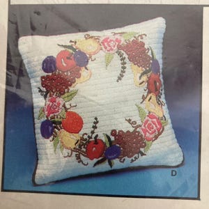 May include: A square, light blue pillow with a detailed embroidered design of a fruit wreath. The wreath includes red apples, purple plums, yellow pears, and pink roses. The pillow has a ribbed texture.