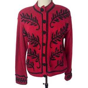 May include: A red cardigan sweater with black leaf-like embroidery and black buttons. The sweater has a high collar and long sleeves. The embroidery is detailed and covers the front and sleeves of the sweater.