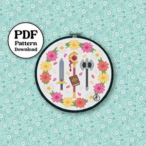 May include: A cross-stitch pattern in a round hoop featuring a floral wreath with pink, yellow, and white flowers. Inside the wreath are a sword, staff, axe, and book. The words "PDF Pattern Download" are in a circle.