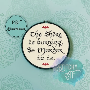 May include: A white embroidery hoop with black stitching around the edge. The hoop has a cream-colored fabric with red and black embroidery that reads "The Shire is burning. So Mordor it is."