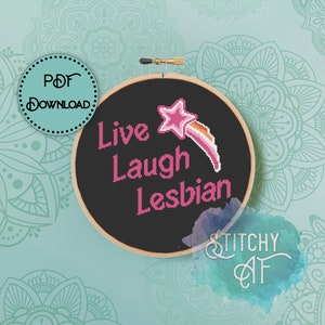 May include: A black embroidery hoop with the text "Live Laugh Lesbian" in pink lettering. A pink star with a rainbow shooting star is above the text. The hoop is on a teal background with mandala designs.