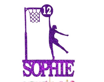 May include: Purple glitter cake topper with a netball player silhouette, a netball, and the name "SOPHIE" and the number "12". The cake topper is on a cake with blue and white frosting and colorful sprinkles. A separate white square cake topper with gold stars and the text "Ooh Let's Party" is on the cake.