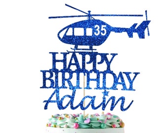 May include: Blue glitter cake topper with a helicopter, the words "Happy Birthday Adam" and the number "35". The cake topper is on a cake with a white square with gold text that says "Ooh Let's Party".
