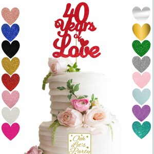 Op de afbeelding: Rode glittertaarttopper met de tekst "40 jaar liefde" en een klein hartje. De topper zit op een witte taart met roze bloemen en de tekst "Ooh Let's Party".