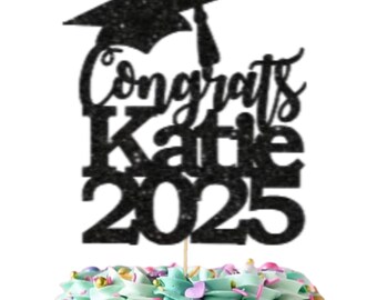 May include: Black glitter graduation cake topper with a graduation cap, the text "Congrats Katie 2025" and a separate white square cake topper with gold stars and the text "Ooh Let's Party".