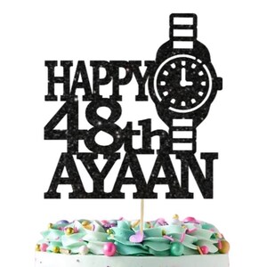 May include: Black glitter cake topper with a watch design and the text "Happy 48th Ayaan". A white square with gold text reads "Ooh Let's Party" on a frosted cake with colorful sprinkles.
