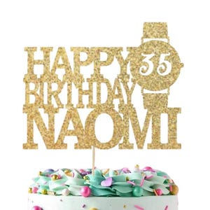 May include: Gold glitter cake topper with the text "Happy Birthday Naomi" and a watch with the number 35 on it. The cake topper is on a cake with blue and white frosting and colorful sprinkles. A separate white square cake topper with gold glitter text says "Ooh Let's Party".