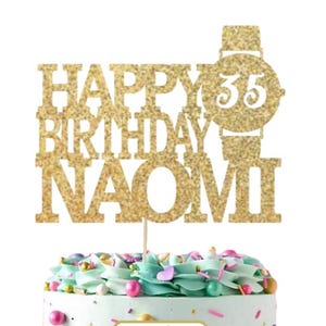 May include: Gold glitter cake topper with the text "Happy Birthday Naomi" and a watch with the number 35 on it. The cake topper is on a cake with blue and white frosting and colorful sprinkles. A separate white square cake topper with gold glitter text says "Ooh Let's Party".