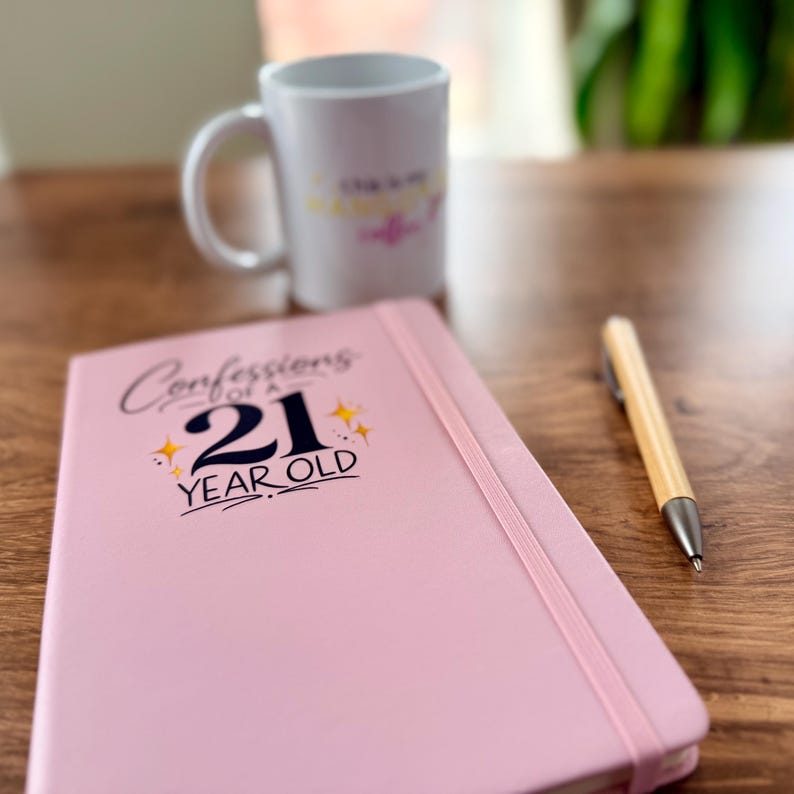 Pode incluir: Um di&aacute;rio rosa com o texto "Confessions of a 21 Year Old" e uma faixa el&aacute;stica rosa a condizer. Uma caneca branca e uma caneta de madeira tamb&eacute;m est&atilde;o na superf&iacute;cie de madeira.