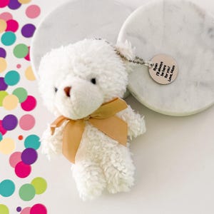 May include: A small, white teddy bear keychain with a tan ribbon bow. The bear has a brown nose and a silver charm that reads "Ryan I'll always be here for you! Love, Mom."