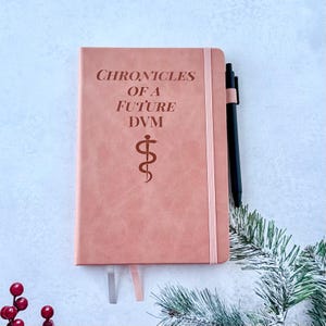 May include: A pink journal with the text "CHRONICLES OF A FUTURE DVM" and a medical symbol. A black pen rests beside the journal. The journal has a pink elastic band and two ribbon bookmarks.
