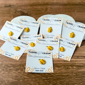 Könnte beinhalten: Eine Sammlung von Baseball-Quastenanhängern. Jeder Anhänger ist ein gelber Baseball mit roten Nähten, der an einer weißen Karte befestigt ist. Die Karten tragen den Text "TASSEL CHARM From Diamonds to Dreams" und "Senior Night 2025-2026".