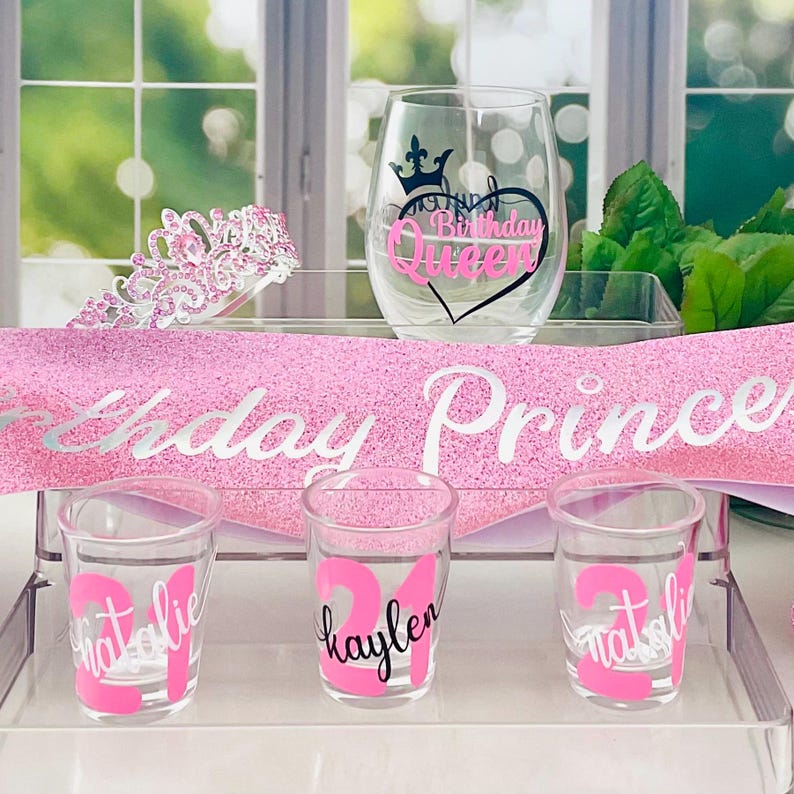 Pode incluir: Um conjunto de decora&ccedil;&otilde;es para festas de anivers&aacute;rio, incluindo uma faixa rosa brilhante com a inscri&ccedil;&atilde;o prateada "Birthday Prince", uma tiara, uma ta&ccedil;a de vinho com o texto "Birthday Queen" e copos de shot com nomes e o n&uacute;mero 21.