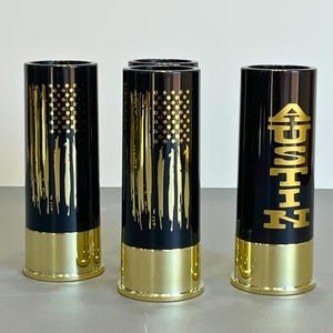 May include: Three black shot glasses with gold accents. Two glasses feature a gold American flag design, and one glass has the name "AUSTIN" in gold lettering.