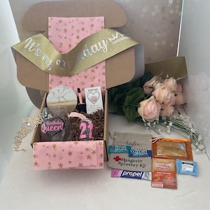 May include: A birthday gift box with a gold ribbon that reads "It's my Birthday". The box contains a tiara, a wine glass with "Birthday Queen", a shot glass with "21", and a bouquet of pink roses. Also included are hangover recovery items.
