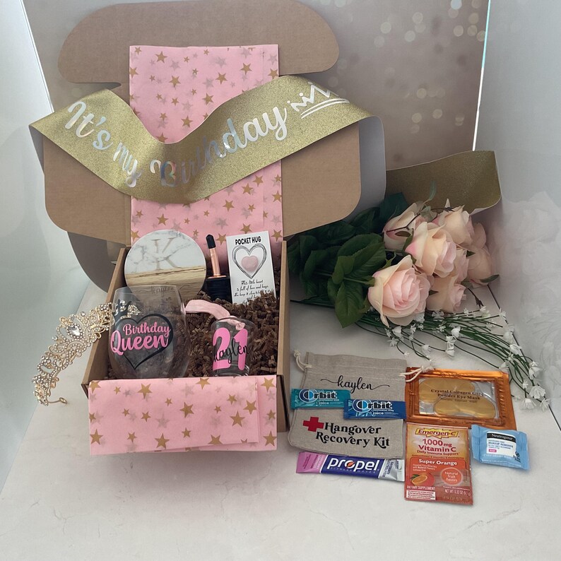 May include: A birthday gift box with a gold ribbon that reads "It's my Birthday". The box contains a tiara, a wine glass with "Birthday Queen", a shot glass with "21", and a bouquet of pink roses. Also included are hangover recovery items.