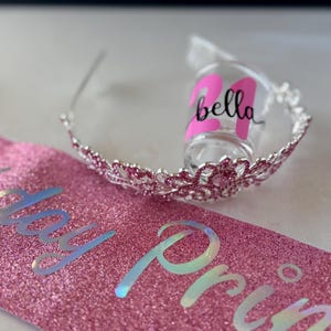 Pode incluir: Uma faixa rosa brilhante com as palavras "Birthday Princess" em letras iridescentes. Uma tiara prateada com pedras preciosas rosa e um copo de shot com o nome "Bella" e o n&uacute;mero "24" em rosa tamb&eacute;m est&atilde;o na imagem.