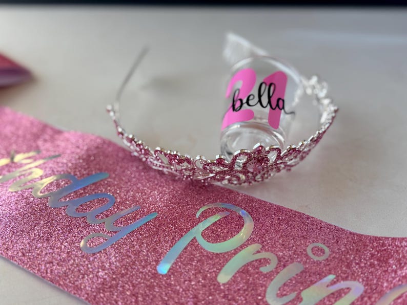 Pode incluir: Uma faixa rosa brilhante com as palavras "Birthday Princess" em letras iridescentes. Uma tiara prateada com pedras preciosas rosa e um copo de shot com o nome "Bella" e o n&uacute;mero "24" em rosa tamb&eacute;m est&atilde;o na imagem.