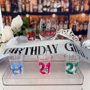 Pode incluir: Uma cole&ccedil;&atilde;o de decora&ccedil;&otilde;es para festas de anivers&aacute;rio, incluindo copos de shot com o n&uacute;mero 21, uma faixa que diz "BIRTHDAY GIRL", uma tiara e uma ta&ccedil;a de vinho com um design de cora&ccedil;&atilde;o e coroa. Os copos de shot s&atilde;o personalizados.
