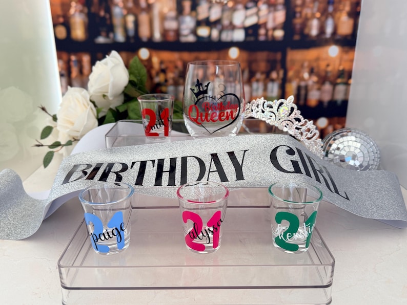 Pode incluir: Uma cole&ccedil;&atilde;o de decora&ccedil;&otilde;es para festas de anivers&aacute;rio, incluindo copos de shot com o n&uacute;mero 21, uma faixa que diz "BIRTHDAY GIRL", uma tiara e uma ta&ccedil;a de vinho com um design de cora&ccedil;&atilde;o e coroa. Os copos de shot s&atilde;o personalizados.