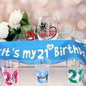 Pode incluir: Uma cole&ccedil;&atilde;o de decora&ccedil;&otilde;es para festas de anivers&aacute;rio. Inclui copos de shot com nomes e o n&uacute;mero 21, uma ta&ccedil;a de vinho com uma coroa e o texto "Birthday Queen", uma faixa azul com o texto "It's my 21st Birthday" e uma tiara.