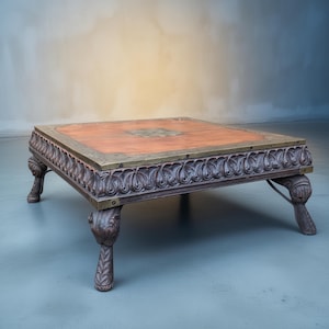 Op de afbeelding: Een vierkante, lage salontafel met een roodbruin houten blad en een sierlijke, donkerbruin gesneden rand. De rand is geaccentueerd met een messingkleurige metalen rand en decoratieve hoekpoten. De tafel heeft een vintage uitstraling.