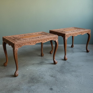 Könnte beinhalten: Zwei antike Beistelltische aus Holz. Jeder Tisch hat eine rechteckige Oberseite mit aufwendig geschnitzten Blumenmustern und geschwungenen Beinen. Die Tische sind warmbraun und stehen auf einem grauen Betonboden.