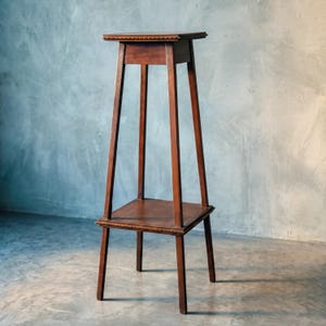 Könnte beinhalten: Ein hoher, dunkelbrauner Blumenständer aus Holz mit einer quadratischen Ablagefläche oben und einem quadratischen Regalboden darunter. Die Konstruktion ruht auf vier schlanken, geraden Beinen und weist an den Kanten beider Ebenen eine dekorative Perlenverzierung auf.