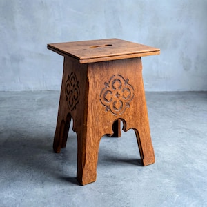 May include: A small, wooden stool with a square seat and carved decorative patterns on the sides. The stool is made of a warm-toned wood and has a cutout in the center of the seat. The legs are angled and have a decorative cutout.