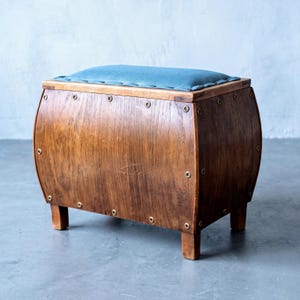 Könnte beinhalten: Ein rechteckiger Fußhocker aus Holz mit einem gewölbten, fassartigen Korpus und einem gepolsterten, blaugrünen Sitz. Das Holz hat eine natürliche Maserung und ist an den Kanten und am Boden mit metallischen, messingfarbenen Ösen verziert.