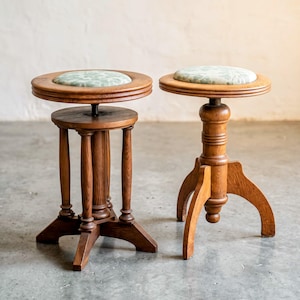 Op de afbeelding: Twee houten krukken met in hoogte verstelbare mechanismen en ronde zittingen bekleed met een lichtgroene stof met een subtiel bloemenpatroon. De krukken hebben gedraaide houten poten en een warme bruine afwerking van de houtnerf.