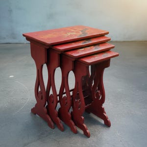 Puede incluir: Juego de cuatro mesas nido antiguas. Las mesas están pintadas de un rojo vibrante con patas curvas decorativas. La superficie de la mesa presenta un diseño ornamentado descolorido en oro y negro. Las mesas están dispuestas en configuración anidada.