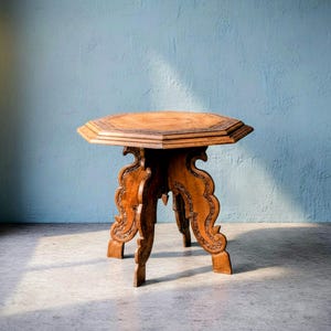 Puede incluir: Una mesa auxiliar de madera vintage con una tapa octogonal y patas talladas ornamentadas. La mesa está hecha de madera de color marrón claro y se apoya sobre tres patas. La mesa probablemente se utiliza con fines decorativos.