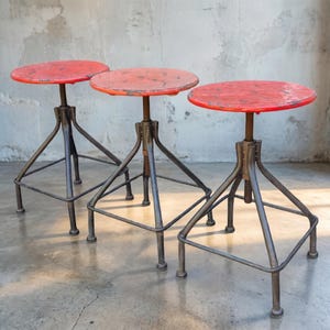 May include: Three industrial-style adjustable stools. Each stool has a round, distressed red seat and a dark metal base. The bases feature a tripod design with a square footrest. The stools are set against a neutral, textured wall.