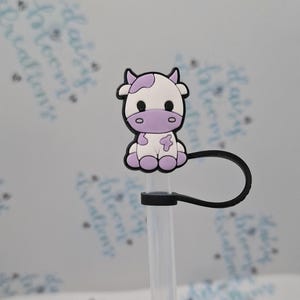 May include: A purple and white cow-shaped silicone straw topper with a black silicone strap. The straw topper is attached to a clear plastic straw.