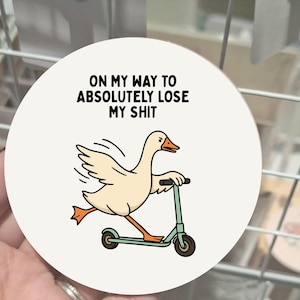 May include: A circular coaster featuring a cartoon goose on a teal scooter. The goose is cream-coloured with orange feet and beak. The text above the goose reads, "ON MY WAY TO ABSOLUTELY LOSE MY SHIT."