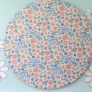 Puede incluir: Un diseño floral redondo con un patrón de flores y lazos rosas, amarillos y azules. Varios recortes de margaritas blancas con centros naranjas están dispersos alrededor del diseño sobre un fondo azul claro. Ideal para manualidades o decoración.