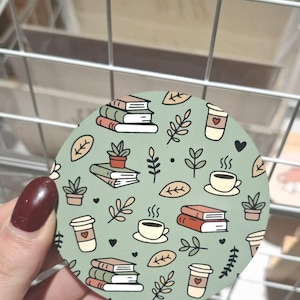 May include: Circular coaster with a green backdrop, showcasing a pattern of books, coffee cups, plants, and leaves. The coaster has a light green background with a repeating pattern of stacked books, coffee cups, and small plants.
