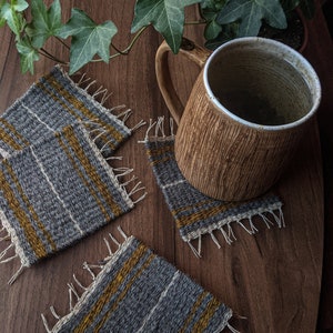Pode incluir: Três tapetes de mesa tecidos com um padrão de listras cinzas e amarelas. Os tapetes têm bordas desfiadas e estão sobre uma superfície de madeira. Uma caneca de cerâmica marrom com uma borda branca está sobre um dos tapetes.