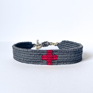 Op de afbeelding: Een grijs geweven armband met een rood kruisontwerp. De armband is gemaakt van een geweven materiaal en heeft een eenvoudig, maar opvallend rood kruis in het midden. De armband is vastgebonden met een natuurlijk gekleurd koord.