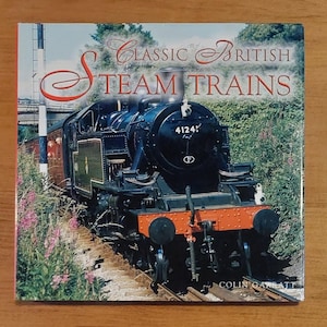 Puede incluir: Portada del libro "Classic British Steam Trains" con una locomotora de vapor negra con frontal naranja. El tren está sobre raíles con follaje verde y cielo azul de fondo. La portada del libro tiene una estética vintage.