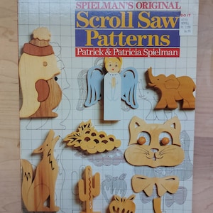 May include: A book titled "Spielman's Original Scroll Saw Patterns" by Patrick & Patricia Spielman. The cover features a grid pattern with various wooden scroll saw patterns, including an angel, a rooster, an elephant, a coyote, a cactus, a grapevine, a cat, a bow, and an apple.