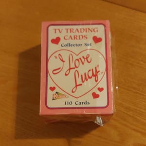 May include: A pink box of "I Love Lucy" TV trading cards. The box features a heart with the show's title and a red heart with a white outline. The box says "Collector Set" and "110 Cards".