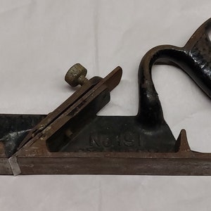 Può includere: Un pialla a mano vintage in metallo nero con base in legno. La pialla ha un manico in metallo e una manopola di regolazione in metallo. La pialla è etichettata "No. 19".