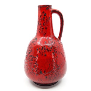 May include: A bright red ceramic pitcher with a bulbous body and a narrow neck. The surface features a textured, mottled black glaze pattern distributed unevenly across the vibrant red base, complemented by a single curved handle on the side.